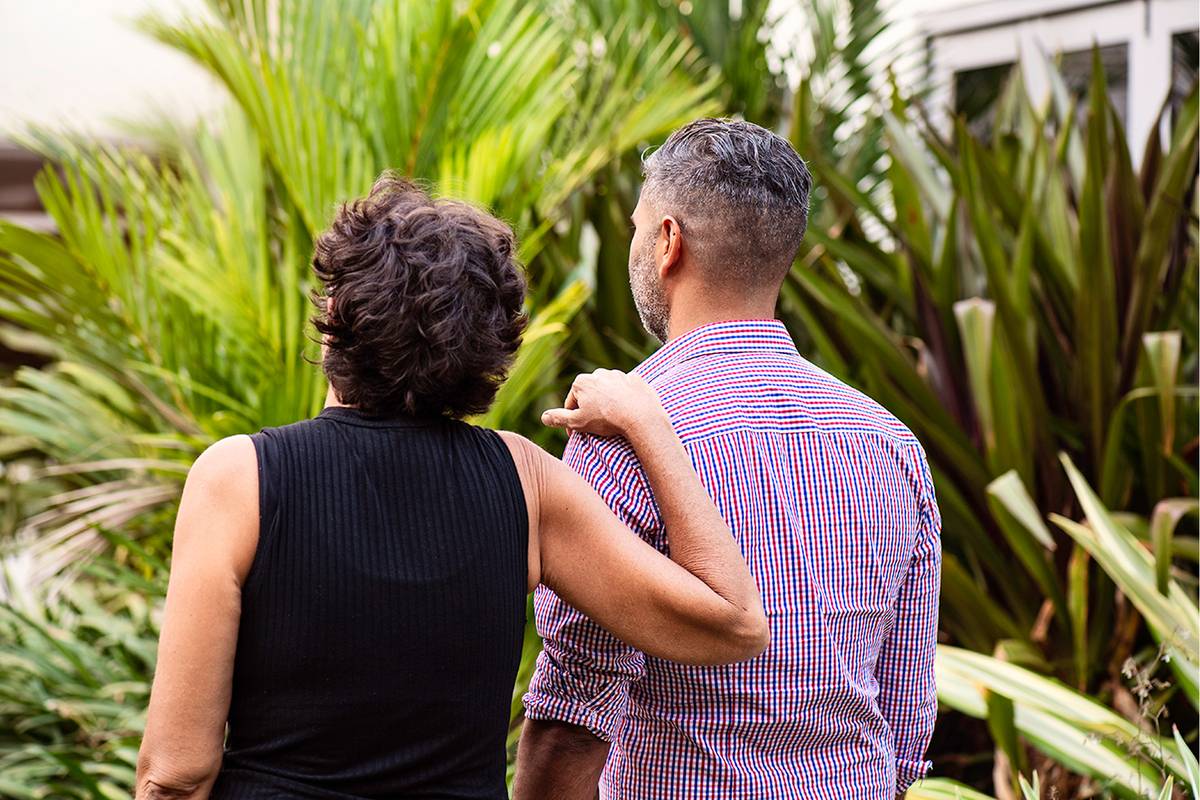 Two people stand together, arm in arm, surrounded by greenery.